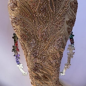 Op de afbeelding: Een lange jurk in goud- en bronskleuren, rijkelijk versierd met kralen en pailletten. Het ingewikkelde ontwerp heeft een vloeiend, organisch patroon. Een decoratief, veelkleurig, geometrisch accessoire is aan de jurk bevestigd.