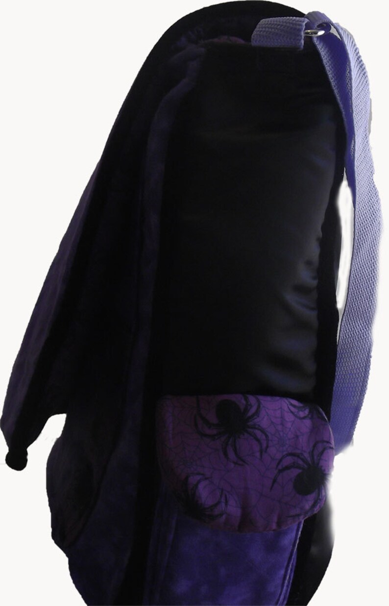 May include: Purple backpack with a black spider web and spider design on the front pocket. The backpack has a purple strap.
