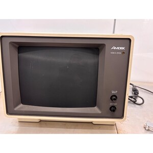 May include: An Amdek Video-310A monochrome computer monitor with a beige plastic casing and a dark gray front bezel. The screen is black, and the front panel features two circular knobs labeled Pull-On Bright and Contrast, plus a power indicator light.
