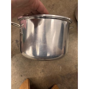 May include: A polished stainless steel stockpot with a cylindrical body, a flat lid, and two side handles. The metal surface reflects light, showing a smooth, metallic finish. The pot features a reinforced rim and a sturdy construction.