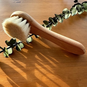 May include: A wooden facial brush with natural bristles. The brush has a long, curved handle and is made of light brown wood. The bristles are white and soft.