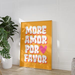 Peut inclure: Une affiche jaune avec le texte "MORE AMOR POR FAVOR" en blanc avec un cœur rose au milieu du texte. L'affiche est encadrée d'un cadre doré.