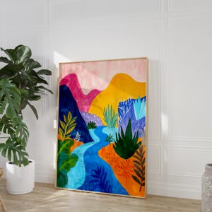 Peut inclure: Une peinture à l'aquarelle encadrée représentant un paysage avec des montagnes, une rivière et un feuillage coloré. Les montagnes sont roses, jaunes et bleues. La rivière est bleue et le feuillage est vert, orange et violet. Le cadre est en bois clair.