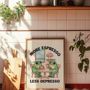 Peut inclure: Une affiche encadrée avec le texte "MORE ESPRESSO LESS DEPRESSO" présente deux grenouilles de dessin animé à une table, chacune tenant une tasse de café. L'affiche est dans un cadre en bois clair et est exposée dans une cuisine.