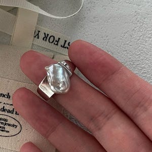 Puede incluir: Un anillo de plata con una perla grande e irregular. La perla es blanca con un ligero brillo iridiscente.