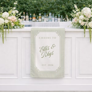 May include: A white bar with a sign that reads "Cheers to Ellis & Rhys EST. 2026" in a decorative frame. The sign has a green and white striped pattern. Floral arrangements with white and pink flowers adorn the bar.