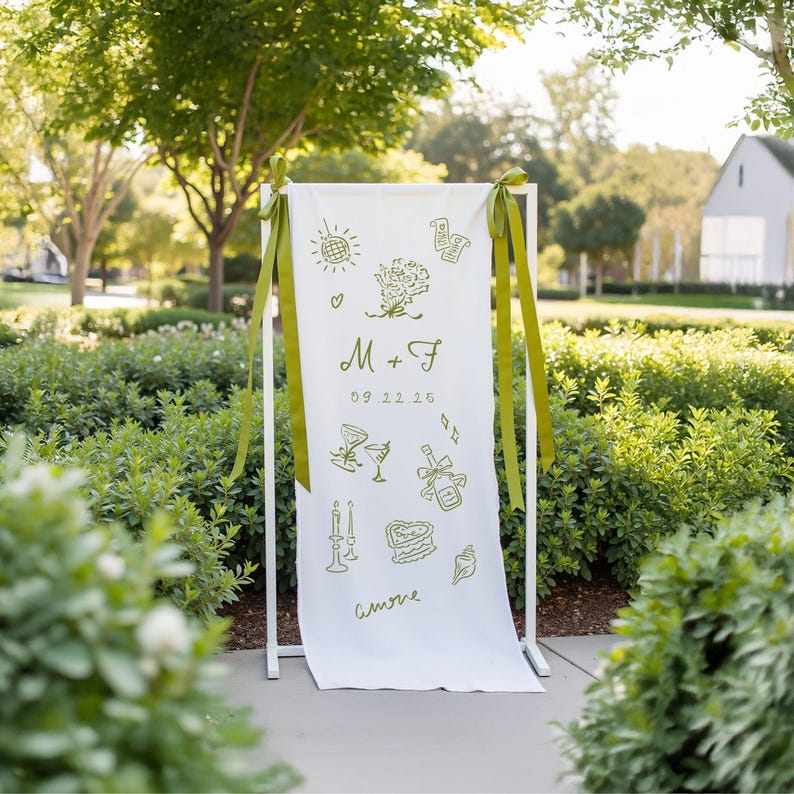 Whimsical fabric wedding sign hanging on a white metal stand, designed with playful olive green line art illustrations such as champagne, wedding cake, bouquet, and candles. The personalized banner includes initials “M + F”, a wedding date, and the word “amore” at the bottom. Perfect for modern, fun-loving couples. Shown in a sunny garden setting with green foliage and ribbon accents.