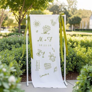 Custom illustrated wedding welcome sign printed on soft white cotton fabric, featuring olive green hand-drawn icons including cocktails, flowers, candles, cake, rings, and a disco ball. The banner includes the couple's initials “M + F” and their wedding date “09.22.25”, and is tied to a white metal frame with matching olive green ribbons. Displayed outdoors with lush greenery in the background.