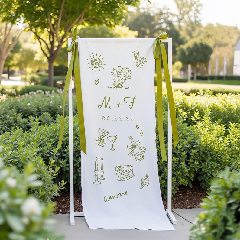 Custom illustrated wedding welcome sign printed on soft white cotton fabric, featuring olive green hand-drawn icons including cocktails, flowers, candles, cake, rings, and a disco ball. The banner includes the couple's initials “M + F” and their wedding date “09.22.25”, and is tied to a white metal frame with matching olive green ribbons. Displayed outdoors with lush greenery in the background.
