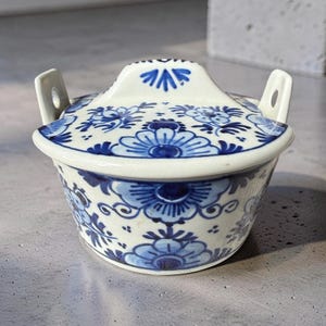 Op de afbeelding: Een kleine, ronde keramische kom met deksel, voorzien van een witte basis met ingewikkelde blauwe bloemmotieven. Het deksel heeft een centrale handgreep en twee zijhandgrepen met kleine ronde uitsparingen. Het ontwerp bevat gestileerde bloemblaadjes.