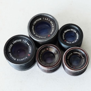 May include: A collection of five vintage camera lenses, each with a dark metal casing and visible glass elements. The lenses are labeled with the brand "CORYGON" and various specifications, including focal lengths and apertures.