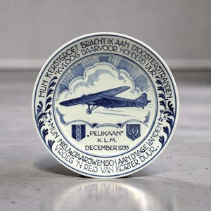 Op de afbeelding: Een wit en blauw keramieken bord met een vliegtuigillustratie, wolken en Nederlandse tekst. Het bord heeft een decoratieve rand en tekst, waaronder "PELIKAAN" en "K.L.M. DECEMBER 1933".