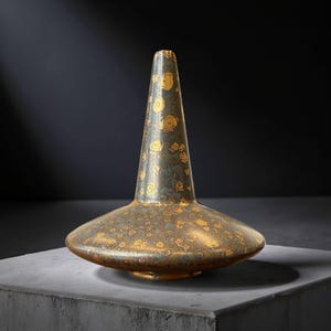Könnte beinhalten: Eine bronzefarbene dekorative Vase mit breitem, flachem Boden und hohem, konischem Hals. Die Vase ist mit einem goldenen Blumenmuster verziert. Die Vase wird auf einer grauen Oberfläche vor dunklem Hintergrund präsentiert.