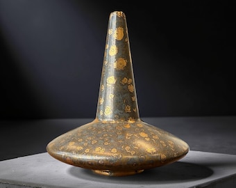 May include: A bronze-colored decorative vase with a wide, flat base and a tall, conical neck. The vase is adorned with a gold floral pattern. The vase is displayed on a gray surface against a dark background.