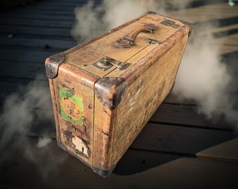 May include: An antique wooden suitcase with metal corner protectors and a leather handle. The suitcase has a worn appearance with visible labels and is surrounded by a hazy, smoky effect, suggesting a journey or travel theme.