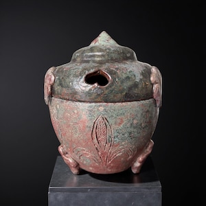 Könnte beinhalten: Ein verwitterter Keramikbehälter im antiken Stil mit Deckel. Der Behälter ist überwiegend rotbraun mit grünen Akzenten und einer herzförmigen Öffnung im Deckel. Der Sockel weist ein geschnitztes Blumendesign auf.