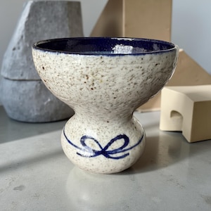 Puede incluir: Un cuenco de cerámica moteada con interior azul oscuro y un diseño decorativo de lazo azul. El cuenco tiene una forma única de reloj de arena y probablemente está hecho a mano. El exterior es de color blanquecino moteado.