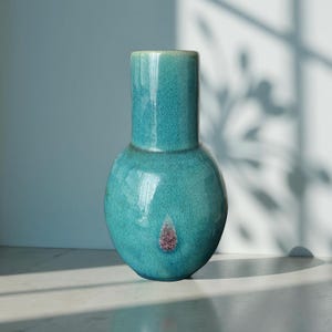 Pode incluir: Um vaso de cerâmica turquesa com base bulbosa e pescoço cilíndrico. O vaso tem um esmalte rachado e um pequeno detalhe rosa-marrom texturizado perto da base. É um item decorativo para casa.
