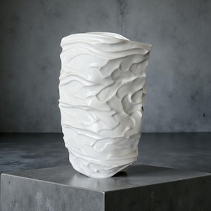 Op de afbeelding: Een witte keramische vaas met een getextureerd, gelaagd ontwerp. De vaas heeft een rechthoekige vorm met een enigszins onregelmatige bovenkant. Het oppervlak heeft horizontale, golfachtige patronen. De vaas staat op een grijze betonnen sokkel.