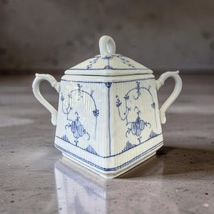 Op de afbeelding: Een witte porseleinen suikerpot met deksel, versierd met blauwe bloemen- en lijnpatronen. De pot heeft twee handvatten en een kleine lus op het deksel. Het ontwerp is symmetrisch en klassiek, geschikt voor het serveren van suiker.