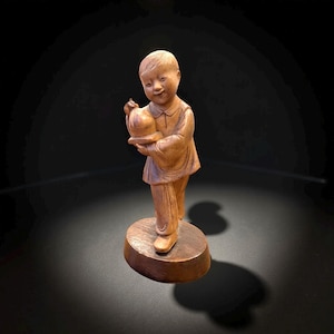 May include: A wooden figurine of a smiling child holding a round object. The figure is carved from light brown wood and stands on a circular base. The child is wearing a long-sleeved shirt.