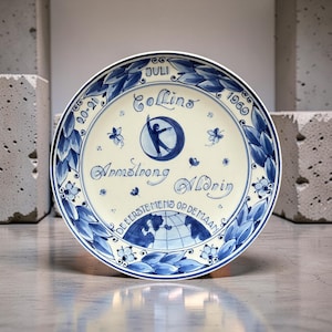 May include: A white ceramic plate with blue hand-painted details. The plate features text and illustrations commemorating the first moon landing. The rim is decorated with blue leaves and floral patterns. The plate includes the names Armstrong, Aldrin, and Collins.
