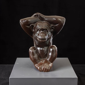Puede incluir: Figura de chimpancé de cerámica marrón con los brazos levantados sobre la cabeza. El chimpancé tiene una expresión sonriente y está sentado sobre una superficie gris. La figura es un objeto decorativo.