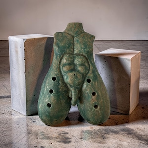 Könnte beinhalten: Grüne Skulptur eines männlichen Torsos mit verwittertem Finish. Die Skulptur hat eine detaillierte Darstellung von Brust und Bauch, mit mehreren kreisförmigen Löchern. Zwei graue Betonblöcke flankieren die Skulptur.