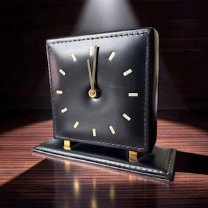 May include: A square black desk clock featuring a smooth leather-like finish with visible stitching along the edges. The clock face displays gold-colored hour markers and thin gold hands, mounted on a rectangular black base with gold supports.
