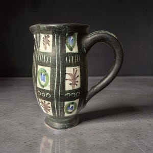 Könnte beinhalten: Eine handgefertigte Keramiktasse mit dunkler, strukturierter Oberfläche. Die Tasse hat einen Henkel und ist mit einem Gittermuster aus weißen Linien und farbenfrohen botanischen Designs in Grün, Blau und Braun verziert. Die Tasse hat einen Fuß.