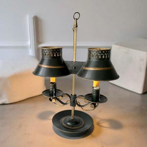 Op de afbeelding: Een vintage kandelaarlamp met twee donkergroene tinten met goudkleurige randen. De lamp heeft twee kaarshouders en een centrale messing steun. De tinten hebben een decoratief geperforeerd ontwerp aan de bovenkant en de basis is bijpassend donkergroen.