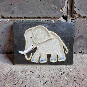 Op de afbeelding: Een rechthoekige keramische tegel met een verhoogd olifantenontwerp. De olifant is wit met een getextureerd oppervlak, omlijnd in lichtbruin en heeft blauwe accenten op de voeten. De tegel heeft een donkergrijze achtergrond.