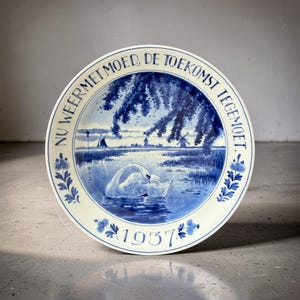 Op de afbeelding: Een vintage blauw-wit keramiek bord met een zwaan op een meer, met windmolens en een zeilboot op de achtergrond. Het bord heeft tekst rond de rand en het jaartal 1937. Bloemen- en bladmotieven sieren de rand.
