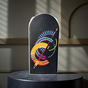 May include: A black ceramic vase with a white rim and a colourful geometric design. The design includes orange, blue, red, and yellow shapes. The vase is on a dark surface.