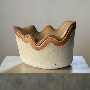 以下が含まれることがあります： 波状の縁とニュートラルな色使いのセラミックボウル。外側は明るいベージュで、縁の周りに赤茶色の帯があります。ボウルは灰色の表面に置かれています。