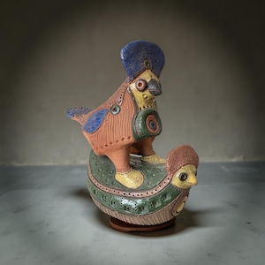 Può includere: Una scultura in ceramica stravagante di due uccelli, uno sopra l'altro. L'uccello superiore ha un copricapo blu e un corpo arancione con accenti blu e gialli. L'uccello inferiore è verde e giallo. La scultura è su una base marrone.