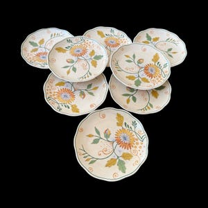 Op de afbeelding: Een set van acht crèmekleurige keramische borden met geschulpte randen. Elk bord heeft een handgeschilderd bloemmotief met oranje, gele en blauwe bloemen, groene bladeren en bruine krullen. De borden staan op een zwarte achtergrond.
