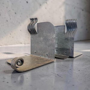 May include: A pair of weathered metal bookends. Each bookend has a rectangular base and a curved, scroll-like design on one end. The top features a series of curved, wave-like accents. The metal has a distressed, aged appearance.