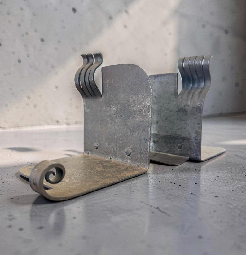 May include: A pair of weathered metal bookends. Each bookend has a rectangular base and a curved, scroll-like design on one end. The top features a series of curved, wave-like accents. The metal has a distressed, aged appearance.