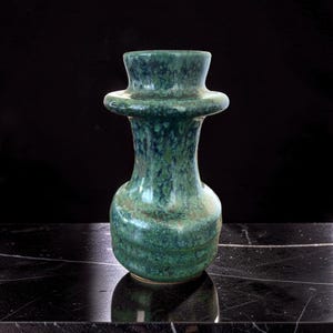 May include: A handcrafted, green ceramic vase with a unique shape. The vase features a wide, flared top, a bulbous midsection, and a narrow base. The surface has a speckled, textured appearance with shades of green and blue.