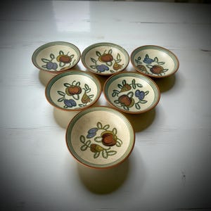 Op de afbeelding: Set van zes kleine, ronde keramische kommen. Elke kom heeft een handgeschilderd ontwerp van fruit en bladeren in bruin, paars en groen. De kommen hebben een crèmekleurige binnenkant en een bruine rand met een groene streep.