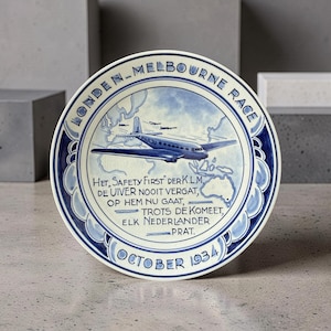 May include: A blue and white decorative plate commemorating the London-Melbourne Race. The plate features an illustration of an airplane over a map, with text in Dutch and the date "October 1934".