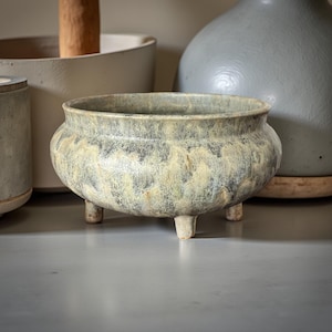 Puede incluir: Un cuenco de cerámica con una superficie texturizada y moteada en tonos grises y crema, apoyado sobre tres pequeñas patas. El cuenco tiene una forma redondeada con una amplia abertura. Otros objetos de cerámica están en el fondo.