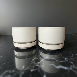 May include: Two cream-coloured ceramic plant pots with matching saucers. The cylindrical pots sit on a flat, round base. The planters are a neutral colour and have a minimalist design, suitable for various plants and home decor styles.