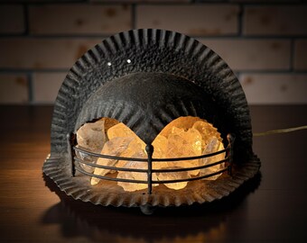 May include: A black metal lamp with a decorative, curved top and a wire cage. The cage holds illuminated, amber-colored crystals. The lamp is on a dark surface, with a brick wall in the background.