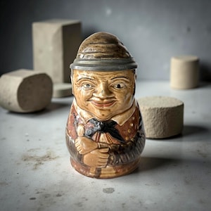 Può includere: Statuetta in ceramica vintage di un uomo con espressione gioviale, cappello e abito marrone e beige dettagliato. La statuetta ha un volto dettagliato e tiene le braccia incrociate. Lo sfondo è neutro con forme geometriche.