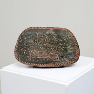 Op de afbeelding: Een keramische schaal met een tankontwerp. De schaal heeft een gedempte kleurenpalet van bruin, groen en grijs. De tank is in het oppervlak geëtst, met een getextureerde rand. De schaal wordt op een wit oppervlak gepresenteerd.