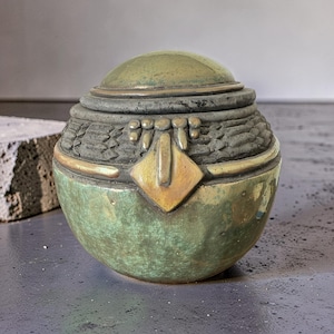 Op de afbeelding: Een decoratieve pot met deksel, met een afgeronde basis en een koepelvormige bovenkant. De pot heeft een getextureerde, donkergrijze band met gouden accenten en een ruitvormige gouden versiering. De basis is een gevlekte groene kleur.