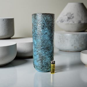 May include: A tall, cylindrical vase with a textured surface and a blend of blue and black colours. The vase is next to a yellow lighter. Other ceramic pieces are in the background.