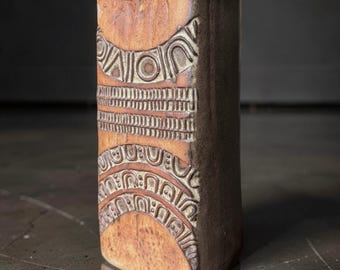 May include: A rectangular ceramic vase with a warm, earthy brown glaze. The front surface features deeply incised geometric patterns, including horizontal bands of repeating arches, circles, and vertical lines, creating a textured, tactile relief.